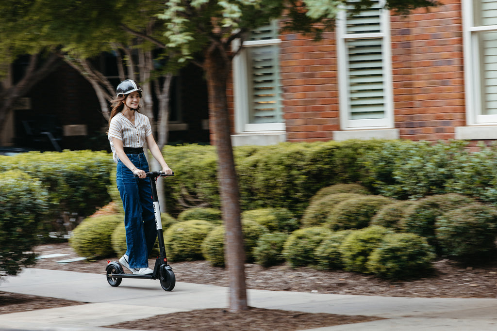 A woman riding the GOTRAX GXL V2 Lightweight Electric Scooter down a campus sidewalk.